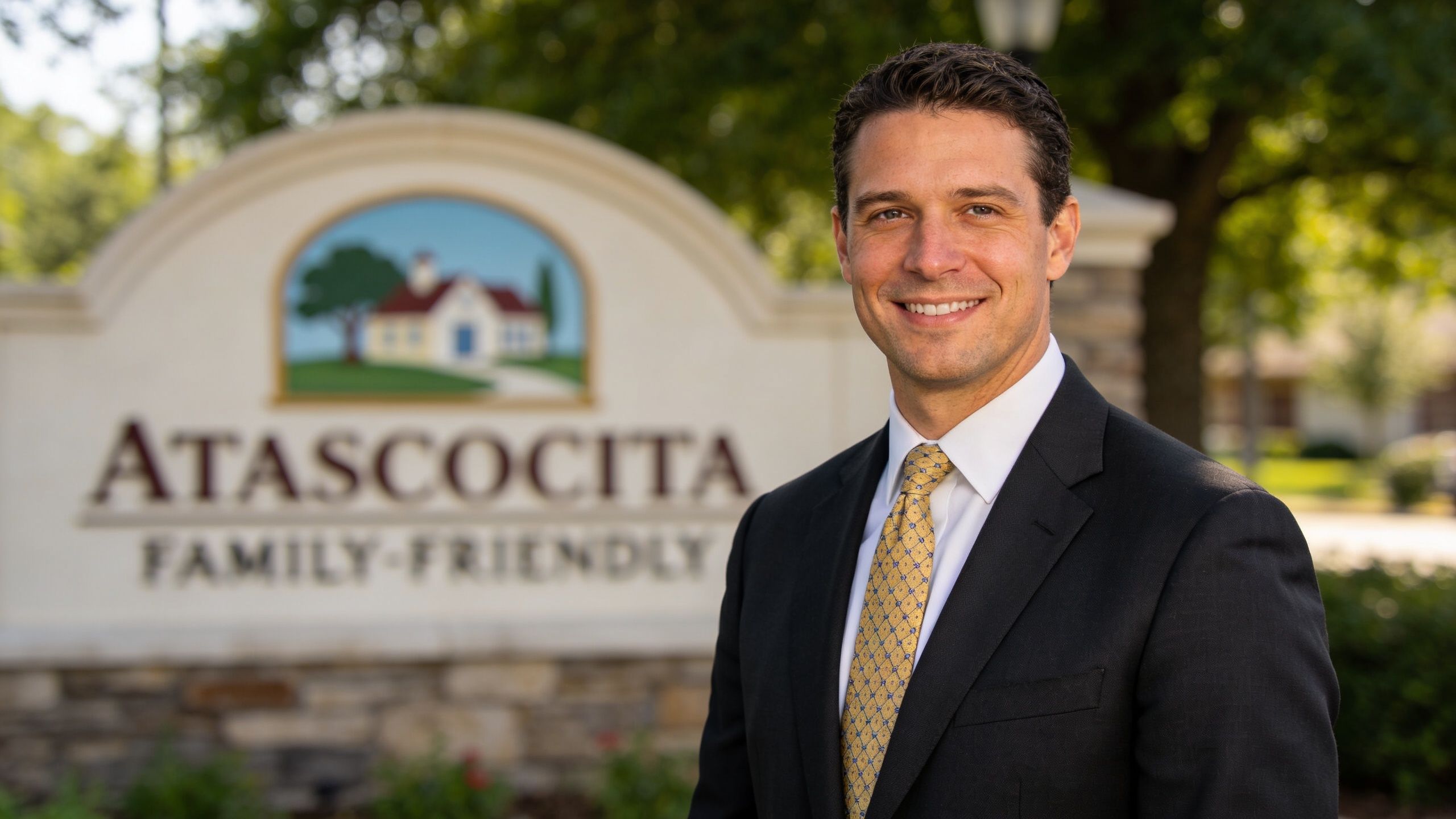 A professional man in a suit and yellow tie standing in front of an Atascocita community sign.