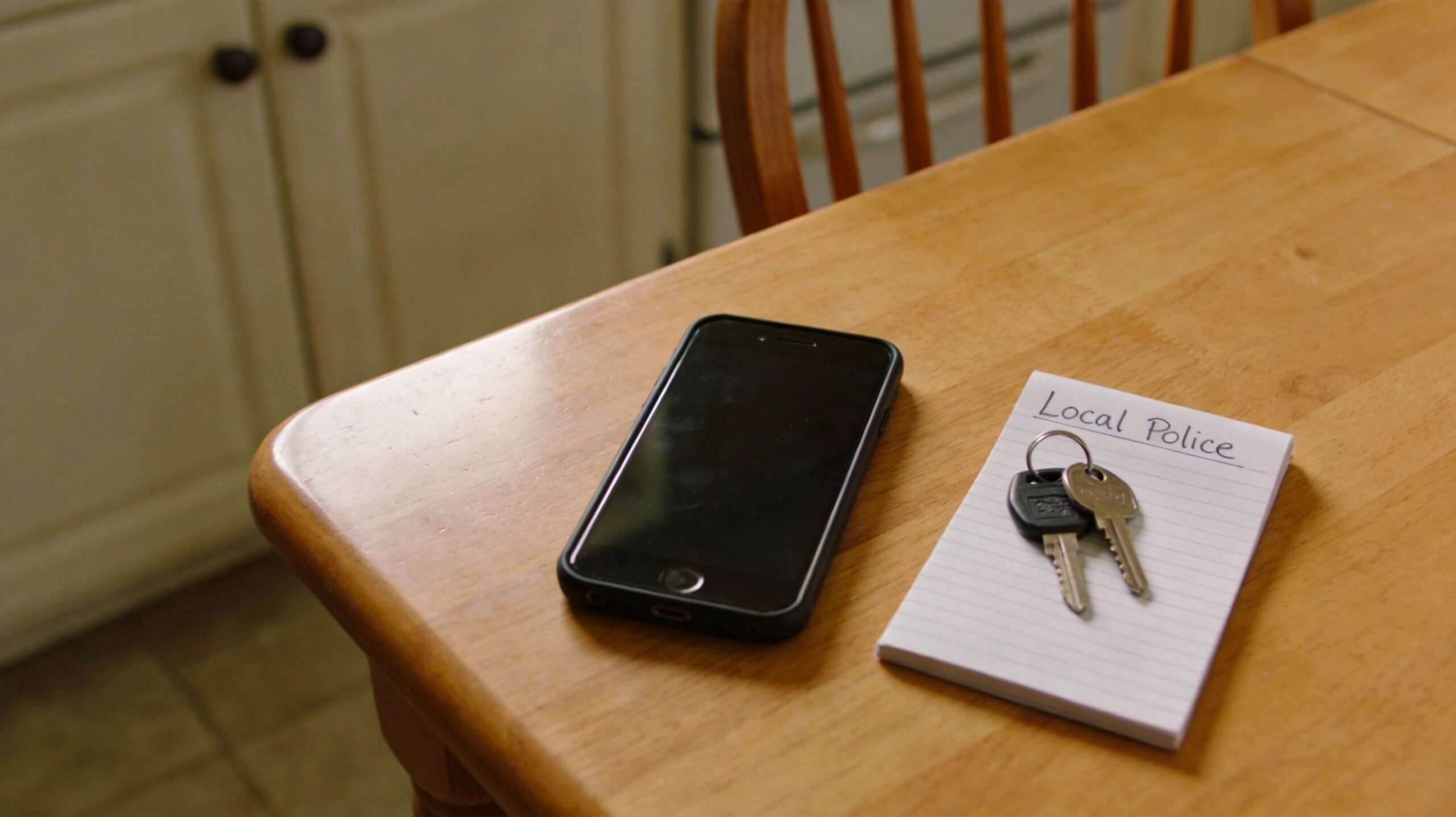 A smartphone and a notepad labeled Local Police with keys resting on a wooden kitchen table.