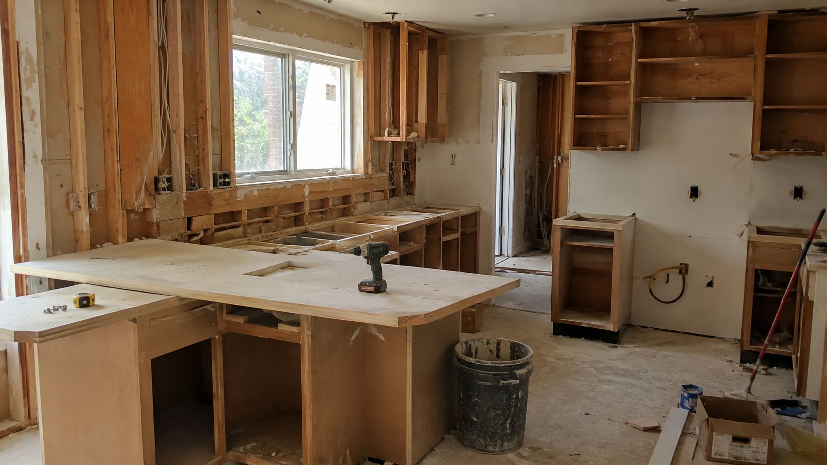 A kitchen under renovation showing unfinished wooden cabinets, countertop installation, and exposed wall framing inside a house.